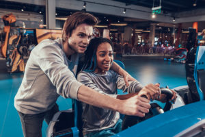 Guy helps his girlfriend drive a fake car in an arcade game. 