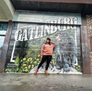 A young woman posing in front of a mural that says "Gatlinburg" with a large black bear on the right. 