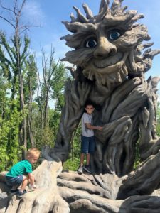 Two kids play around a troll statue that is made to look like he is made of willow trees. 